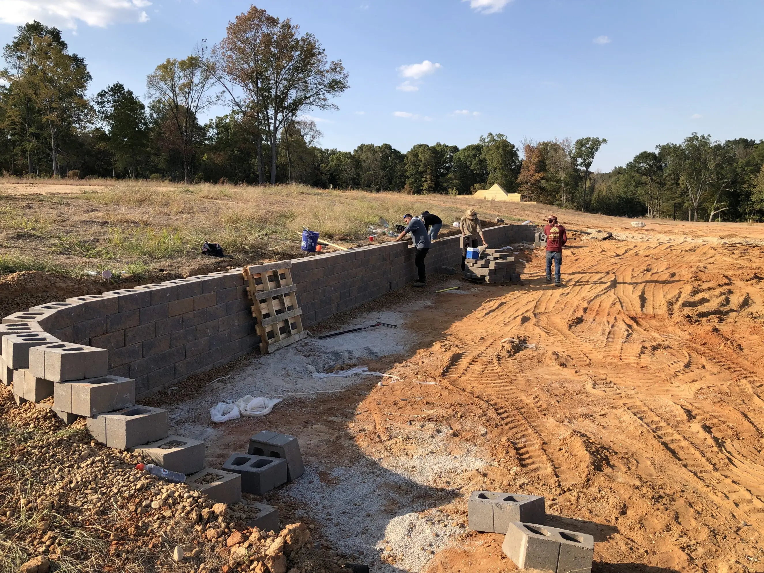 Workers installing a curved retaining wall of stacked concrete blocks on a cleared lot, demonstrating hardscape construction for landscaping services and patio/edging installation.