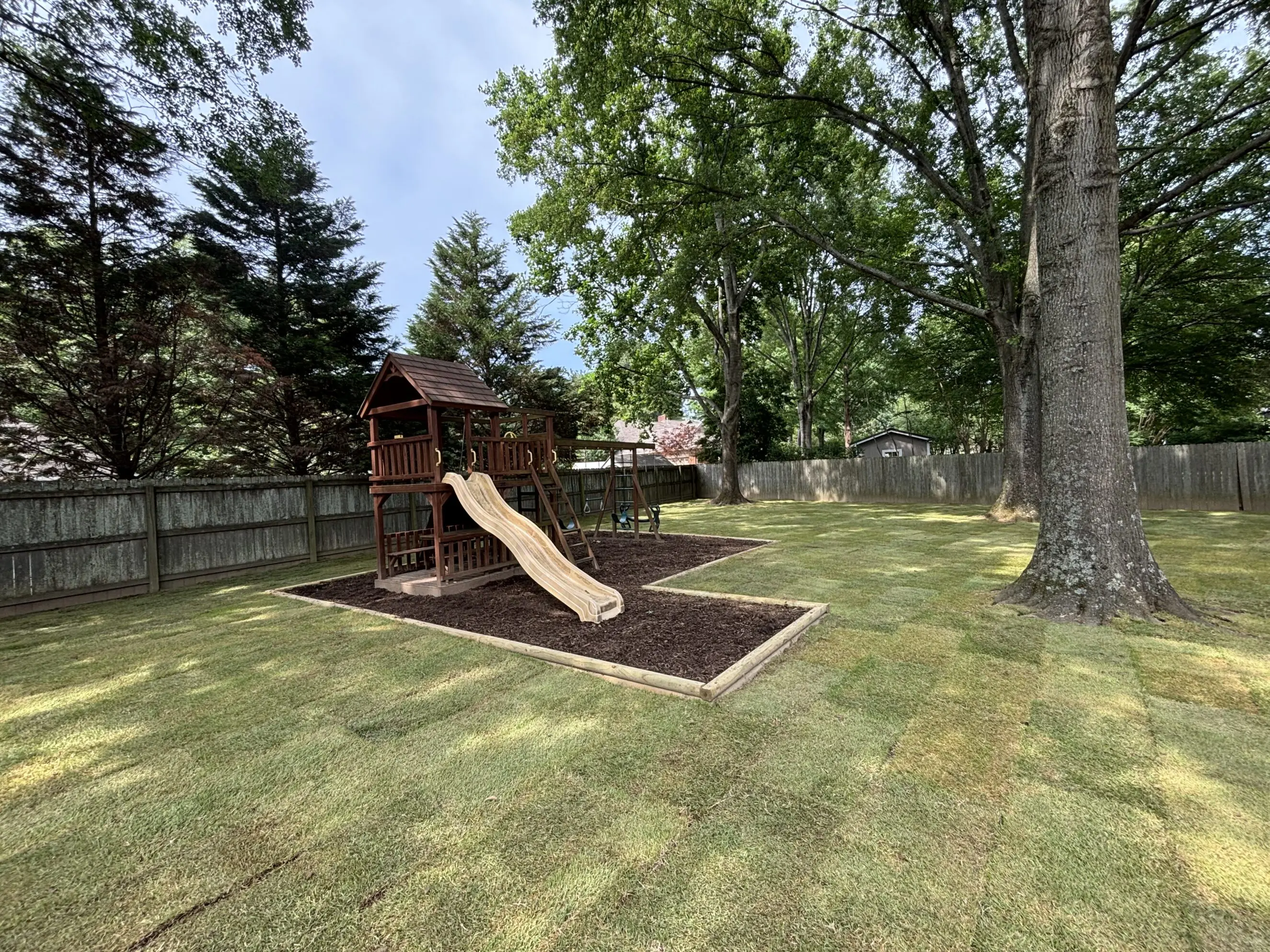 Backyard landscape with fresh sod, bordered mulch play area featuring a wooden playset and slide, mature shade trees, and a fenced perimeter — professional landscaping and lawn installation in a residential yard