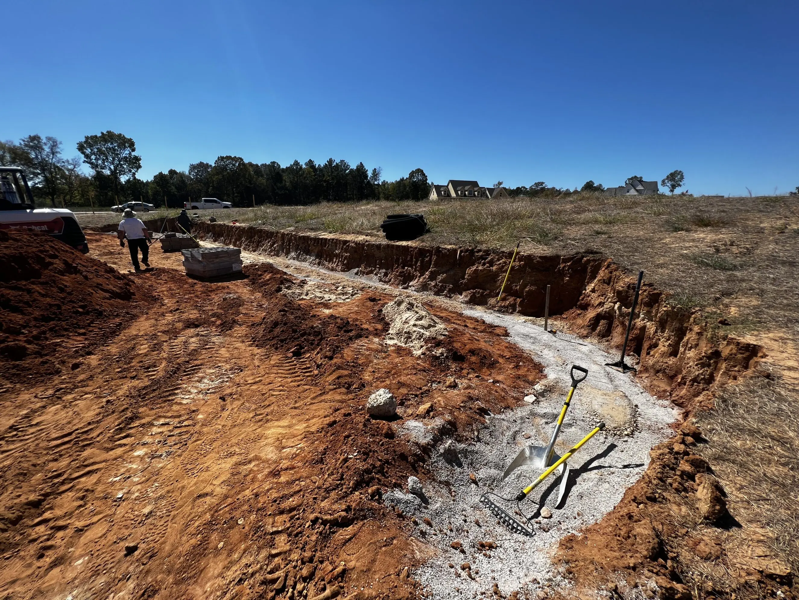 Landscaping crew preparing a trenched garden bed with shovels, rake, gravel base and compacted red clay soil at a residential yard renovation by Bluff City Tree & Landscape in Memphis-area community